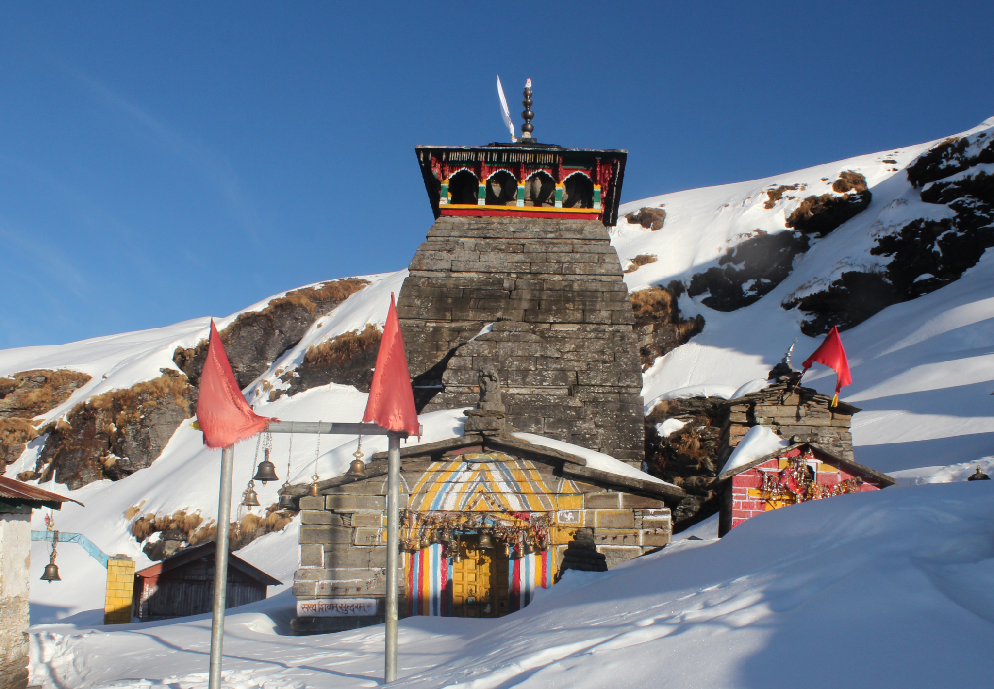 A scenic view of Tungnath Temple, nestled among the lush green mountains, with its intricate stone architecture standing tall under a clear blue sky. The temple is adorned with colorful flags, and the surrounding area is dotted with vibrant wildflowers and verdant vegetation. Snow-capped peaks can be seen in the distance, adding to the temple's serene and majestic ambiance.